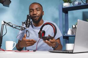 African american blogger showing headphones for giveaway, speaking in microphone and recording video for channel. Young streamer holding headphones, talking in mic and streaming live photo