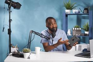 African american blogger reviewing photography equipment and sharing insights with internet channel audience. Vlogger holding camera power accumulator and talking while live streaming photo
