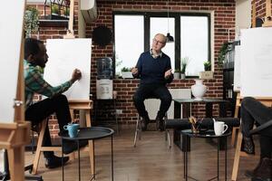 Senior man drawing teacher talking with adult students during group art class. Diverse people sitting at easels engaged in artmaking, learning to draw with tutor, making time for creative hobby photo