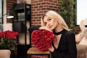 Lovely girlfriend holding flower bouquet in heart shaped box, enjoying celebrating valentine s day. Attractive blonde woman with in black dress standing in living room filled with romantic presents photo