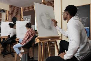 Diverse people engaged in arts activities. Young African American guy practicing sketching skills during art workshop, learning to draw in group, taking up drawing class, creating masterpiece photo