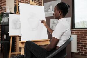 Young focused African American woman learning to draw at group drawing class, sitting at easel in classroom and sketching object with pencil on canvas, people taking up art classes for stress relief photo