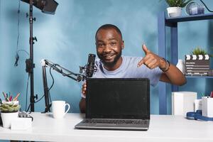 Smiling african american blogger pointing at laptop with black screen while recording product presentation. Vlogger presenting digital device and streaming review on social media photo