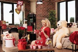 Cute girlfriend sitting in living room filled with romantic gift, enjoying celebrating love holiday. Attractive sexy woman in red dress receiving valentine s day presents from boyfriend photo