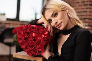 Smiling woman posing with red roses in heart shaped box, celebrating valentine s day in living room filled with romantic presents from boyfriend. Cute girlfriend in black dress smiling at camera photo