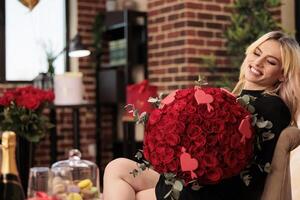 Girlfriend standing in living room filled with romantic gifts, holding red roses large bouquet. Beautiful blonde woman in elegant black dress enjoying celebrating valentine s day. Love concept photo