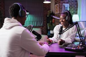 Online show host interviewing african american woman making him laugh during live broadcast. Talking show interviewer using professional microphone and decor to record comedy podcast photo