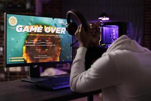 Gamer upset after losing online multiplayer action videogame tourney, being defeated by other players. African american man removing headphones in frustration after receiving game over message photo