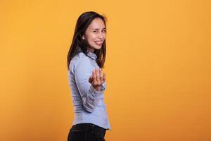 Smiling cheerful asian model doing come to me symbol, beckoning with finger posing in studio over yellow background. Brunette woman inviting you for confidential talk. Calling gesture photo