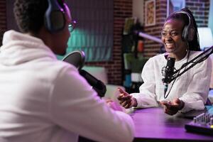 Online show host enjoying conversation with african american woman sharing favourite jokes during comedy focused podcast. Talking show interviewer in living room studio broadcasting interview live photo