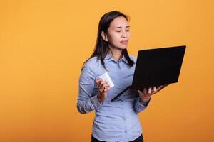 Asian woman introducing credit card information on laptop computer, doing online shopping in studio over yellow background. Cheerful young adult buying fashion clothes via internet transaction photo