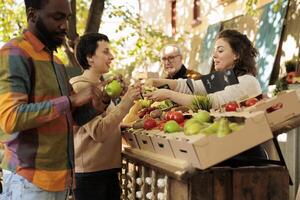 Young diverse family couple tasting fresh organic produce while shopping at outdoor farmers market. Friendly female vendor standing behind farm stall offering customer to taste apple before purchase photo