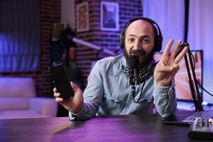 Portrait of tech vlogger presenting three tips for finding smartphones with best value on the market in rgb lights home studio. Content creator filming video, showing counting gesture with fingers photo