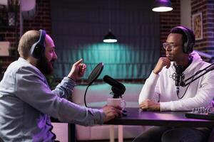 African american influencer recording podcast with guest, using audio recording equipment in home studio. Cheerful men chatting live during broadcasting session for internet show photo