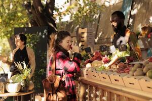 joven mujer disfrutando compras a local agricultores mercado en soleado otoño día. hembra oliendo Fresco maduro manzana con cerrado ojos mientras comprando orgánico comida frutas y verduras, visitando cosecha festival foto