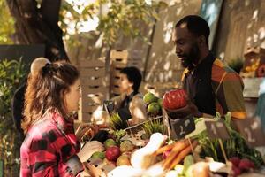 Man vendor standing behind stall selling fresh organic vegetables to female customer, displaying produce at local farmers market. Young woman buying natural home-grown produce photo