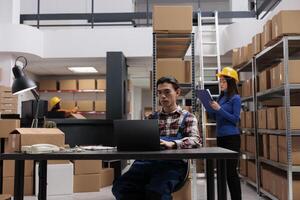 postal Servicio empleado rastreo paquete o empaquetar envío en ordenador portátil mientras sentado a mesa en enviar oficina almacenamiento habitación. asiático entrega empleados haciendo inventario administración en almacén foto