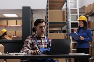 Young asian warehouse employee working on laptop in post office storage room. Delivery service worker analyzing customer orders checklist on computer and managing dispatching photo