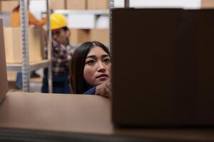 Asian woman warehouse worker doing inventory management while checking customer parcels on shelf. Logistics manager searching cardboard box in distribution center storehouse photo