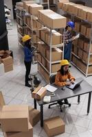 Industrial storehouse asian coworkers doing inventory in storage room full of cardboard boxes on shelves. Postal service warehouse young colleagues team in protective helmets working together top view photo