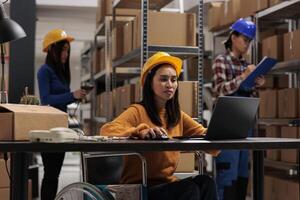 Young asian woman wheelchair user working on laptop in warehouse. Industrial storehouse employee with physical disability checking goods in stock on computer in inclusive workplace photo