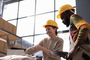 diverso compañeros de trabajo vistiendo industrail en general y casco, mientras comprobación en línea pedidos en ordenador portátil computadora en almacenamiento habitación. almacén equipo analizando entrega detalles en digital dispositivo en almacén foto