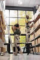 Storehouse workers looking at shelves full with clients orders, carrying cardboard box while listening music at headphones. Employees working at products packages preparing delivery in warehouse photo