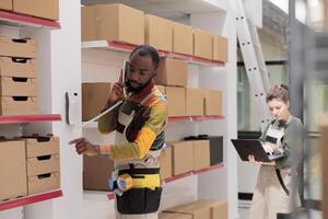 African american manager discussing merchandise logistics with remote supervisor at landline phone in warehouse. Employee working at customers orders, checking shipment details in storehouse photo