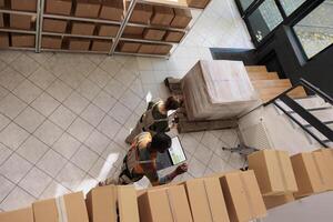 Diverse employees tracking clients online orders, checking warehouse logistics using laptop computer. Storage room supervisor preparing packages for delivery with manager. Top view concept photo