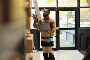 Storage room employee looking at cardboard boxes, checking merchandise checklist on tablet computer before start preparing customers orders. African american supervisor working in warehouse photo