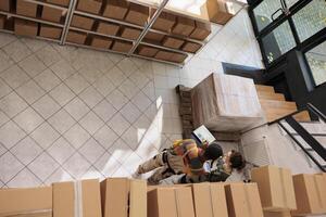 Top view of workers checking customers orders on laptop before packing and shipping, working in storage room. Diverse team weraring protective overall overseeing online production of merchandise. photo