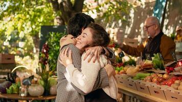 Woman vendor greeting regular client and hugging at farmers market stand, being happy selling locally grown products. Positive farmer sharing hug with customer, enjoying visit at street festival. photo