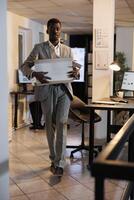 Sad manager carrying cardboard box with belongings after being dismissed from startup office. African american businessman leaving office late at night. Business management concept photo