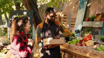 Cheerful farmers market seller putting fresh produce in box, selling package of natural eco fruits and vegetables. Young woman talking to local marketplace vendor, buying organic products. photo