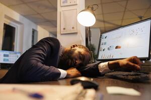 Exhausted tired executive manager suffering from sleepiness after working late at night in startup office. Drained african american worker with burnout syndrome sleeping at work. Business concept photo