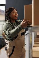 Cheerful manager taking out cardboard box from shelves, preparing customers order during work shift in warehouse. African american employee wearing industrial overall working at products delivery photo