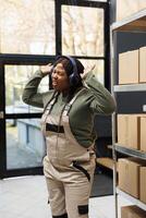 Small business employee dancing during work break in warehouse, listening music and having fun before start working at storage room inventory. African american worker wearing headphones photo