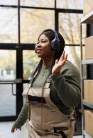 Cheerful manager wearing headphones listening music during work break in storehouse, dancing and having fun. African american employee with industrial overall doing merchandise quality control photo