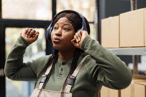 Supervisor with headphones dancing and having fun, working at customers orders preparing packages for shipping in warehouse. Storage room worker in industrial overall listening music photo