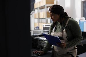 African american manager checking customers online orders on computer before start preparing packages for shipping in storage room. Warehouse employee wearing industrial overall standing at counter photo