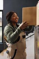 Stockroom worker taking out cardboard box from shelves, preparing customers online orders in warehouse. African american supervisor wearing industrial overall working at products delivery in storehouse photo