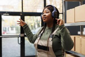 Manager wearing headset and having fun during work break, enjoying free time before start working at customers orders. African american employee listening music and dancing in warehouse photo