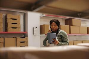 African american supervisor having remote conversation with manager discussing shipping details in warehouse. Employee looking at merchandise checklist on tablet computer in storage room photo