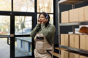 Stockroom supervisor wearing headset listening music, enjoying work break in warehoue. African american employee having fun with song before start preparing customers packages in storehouse photo