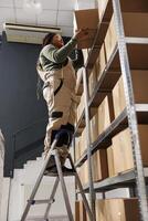 African american employee standing on ladder in storehouse, preparing customers orders using cardboard boxes. Small business worker wearing overall doing merchandise quality control in warehouse photo