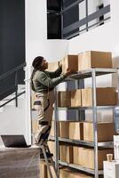 Warehouse worker taking out carton boxes from shelf, preparing clients orders in storage room. African american stockroom manager working at shipping details for packages in storehouse photo