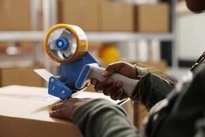 Storehouse worker using adhesive tape to pack clients orders, preparing packages for delivery in storehouse. African american employee wearing industrial overall during merchandise inventory. Close up photo