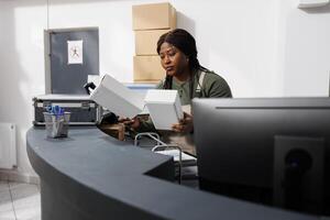 Storehouse employee looking at white carton boxes, checking customers orders on computer in storage room. African american supervisor wearing industrial overall while preparing packages for delivery photo