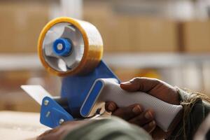 Stockroom worker putting adhesive tape on cardboard boxes, preparing clients orders in warehouse. African american employee checking products before shipping packages in storage room. Close up photo