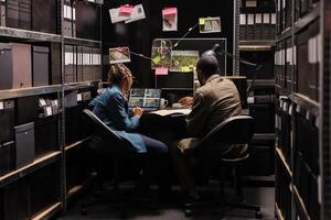 Private detectives looking at surveillance photos on laptop and analyzing report. African american man and woman investigators examining clues in dark agency office at night